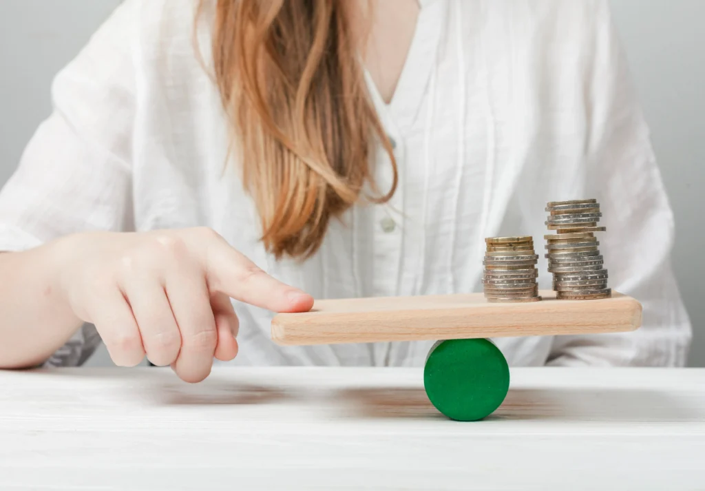 A person balances a wooden plank with stacks of coins on a fulcrum, symbolizing the consideration and balance of cost versus value when deciding on the cost of lash extensions.