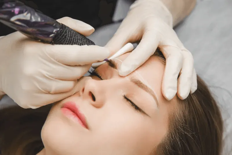 The image shows a close-up of the microblading procedure being performed on a woman's eyebrows,