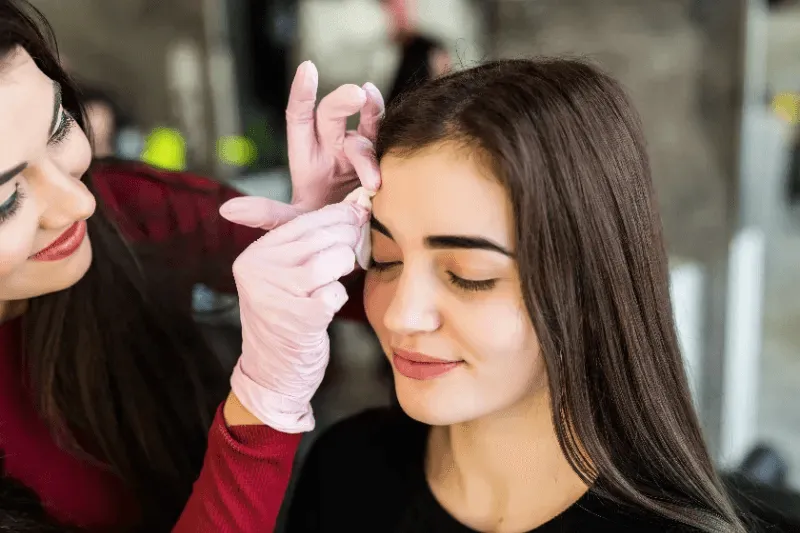 The image shows a practitioner gently cleaning a client's eyebrows, which is part of the aftercare process