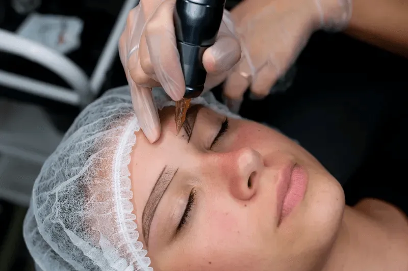 The image shows a professional using a tattooing machine to create precise eyebrow tattoos, highlighting the focus and detail involved in the process.