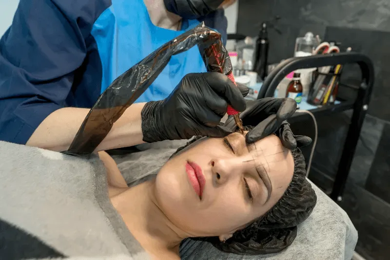 The image depicts a close-up of an eyebrow tattooing procedure in progress