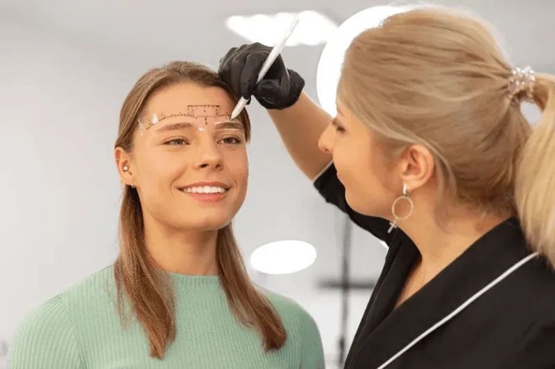 The image shows a professional marking a client's eyebrows with precision tools, symbolizing the initial steps of a permanent eyebrow procedure consultation.